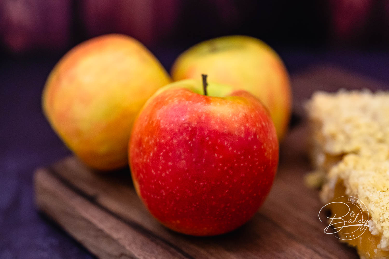 Drei frische Äpfel auf Holzbrett neben einem Apfelkuchen mit Streusel
