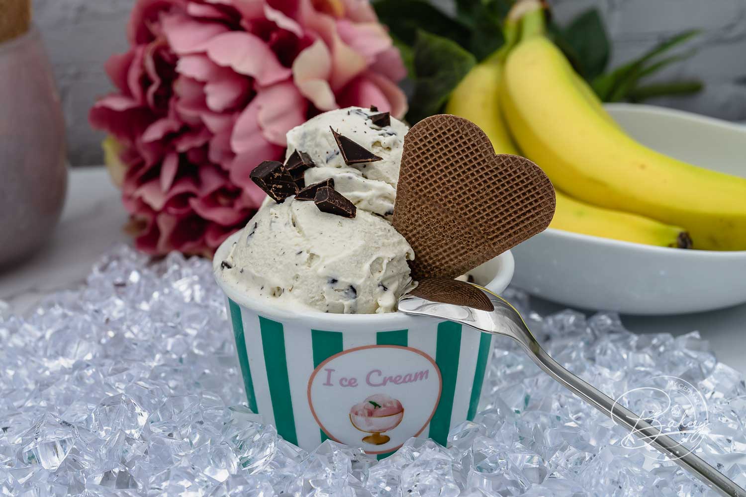 Bananen Eis mit Schokostücken und Waffelherz im Eisbecher, dahinter frische Bananen