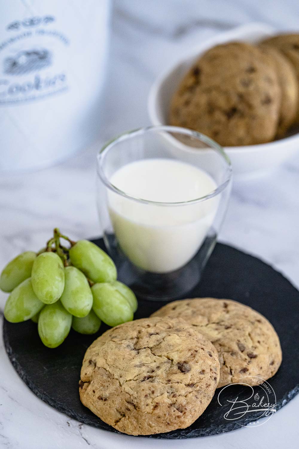 Zwei goldbraune Chocolate Chip Cookies neben grünen Trauben, Milchglas und Keksschale