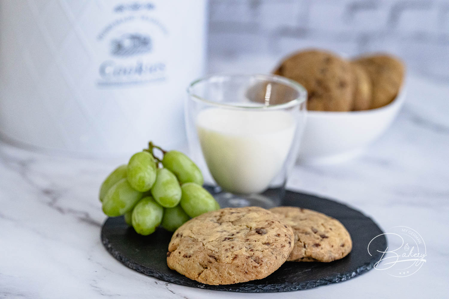 Zwei Chocolate Chip Cookies auf Schieferplatte mit Trauben und einem Glas Milch