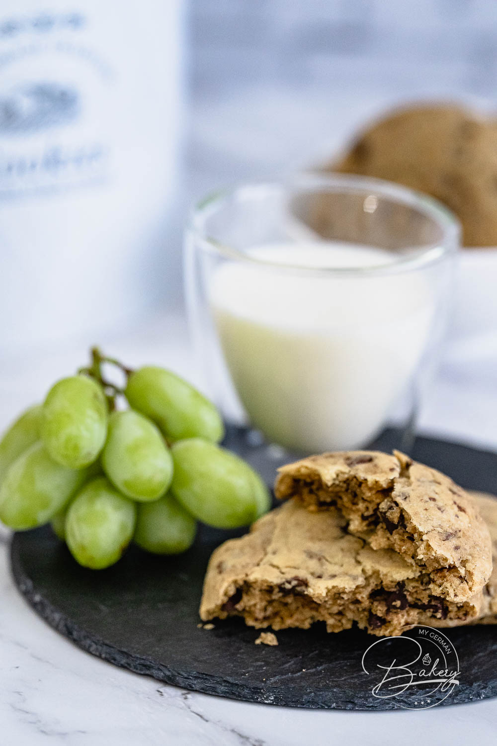 Milchglas vor Keksdose mit Trauben und angebissenem Chocolate Chip Cookie am Bildrand