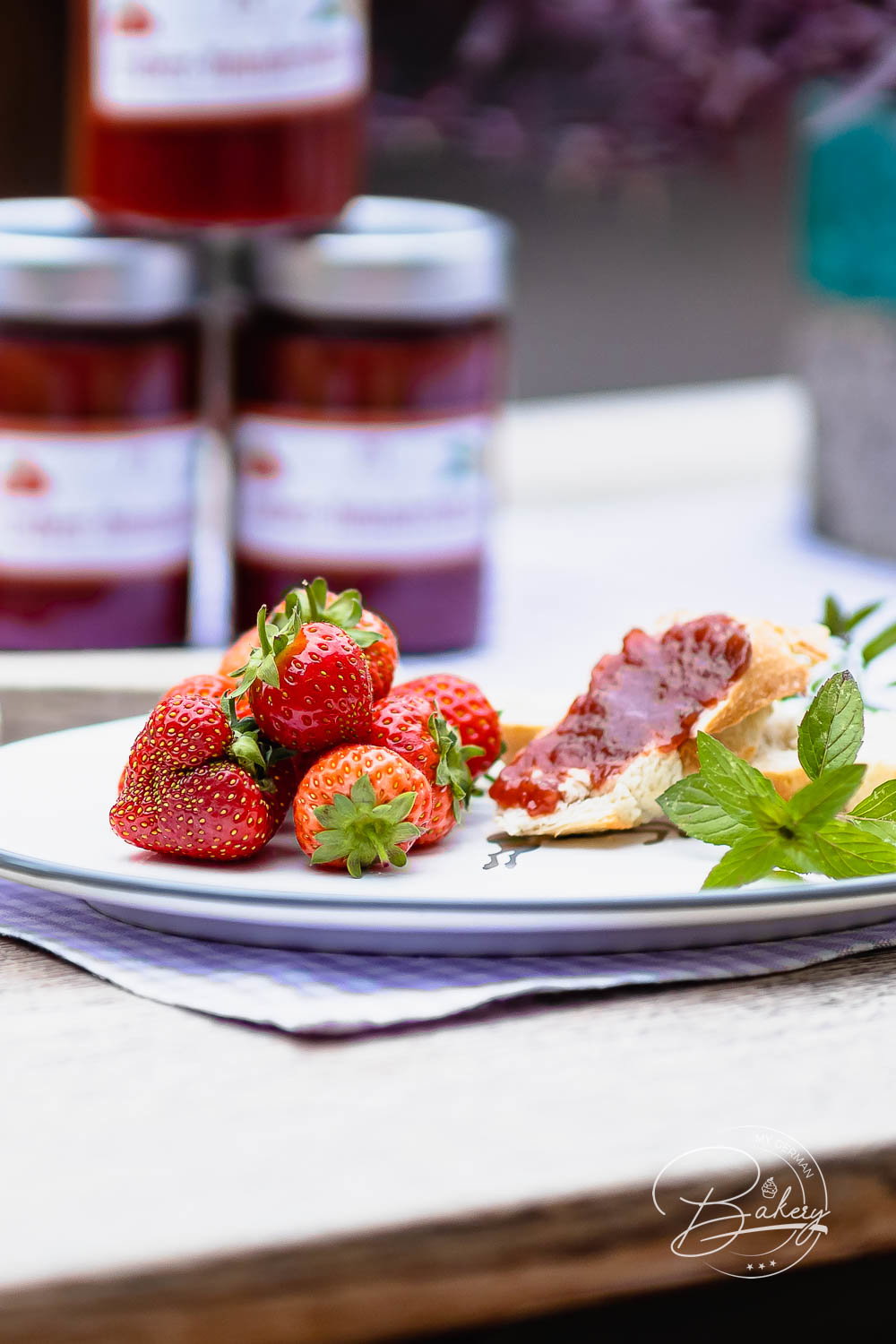 Drei Gläser selbst gekochte Erdbeermarmelade hinter einem Teller mit frischen Beeren