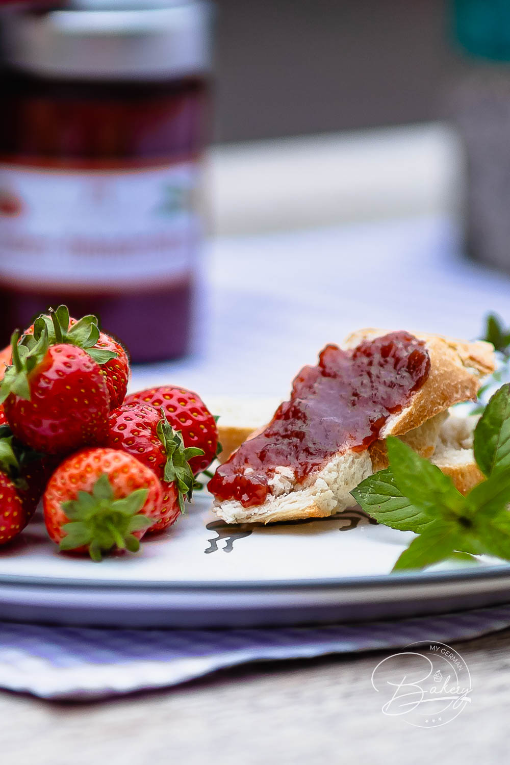 Teller mit Erdbeeren, Minze und Marmeladenbrot vor einem Glas Erdbeermarmelade
