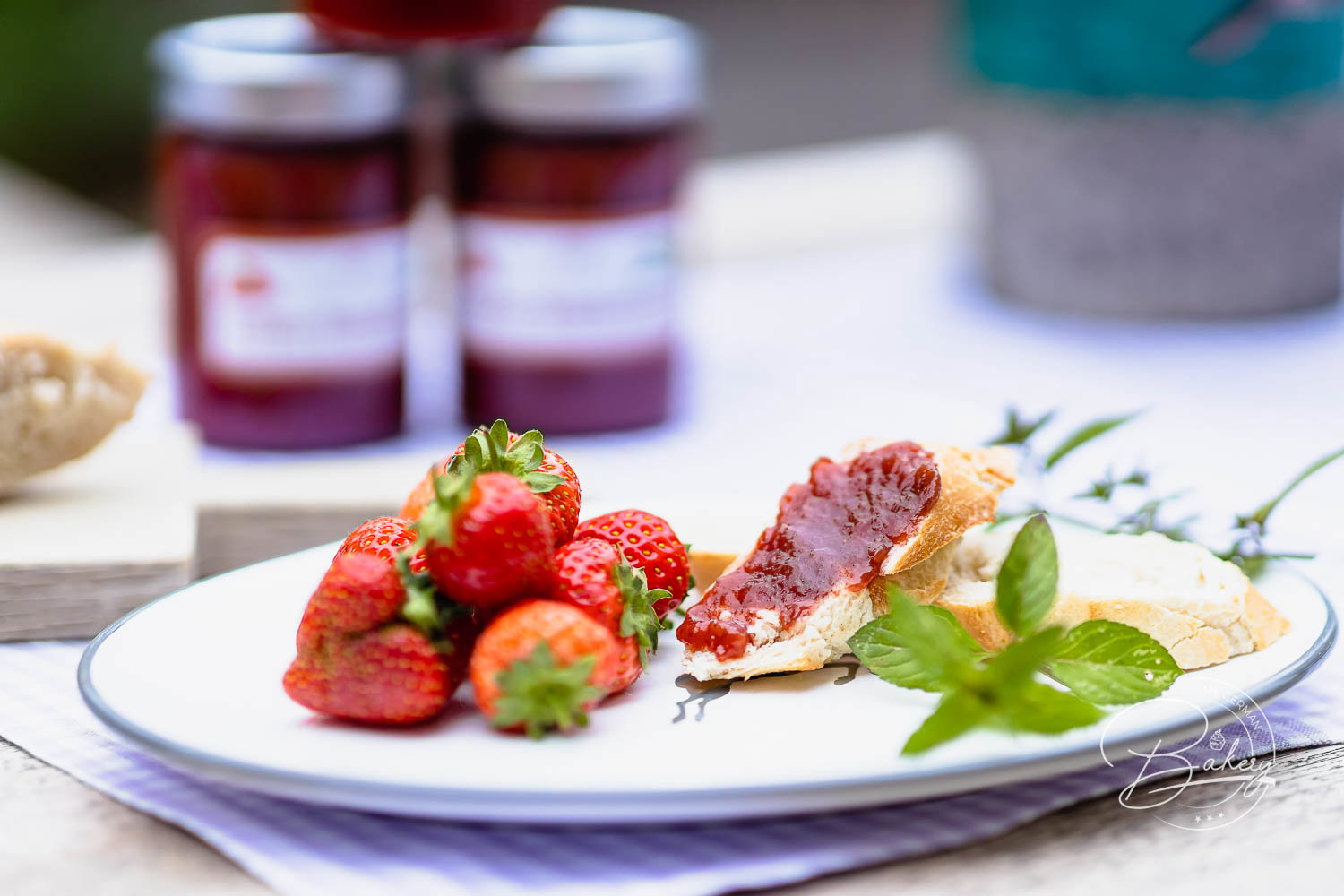 Frühstücksteller mit Erdbeeren, Minze und bestrichenem Baguette neben zwei Gläsern Erdbeermarmelade