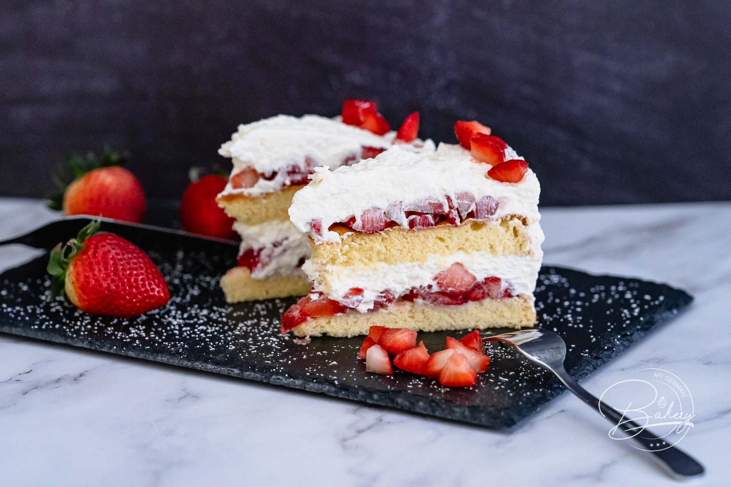 Stück Erdbeer-Sahne-Torte mit zwei Biskuitböden und frischen Erdbeeren auf Schieferplatte