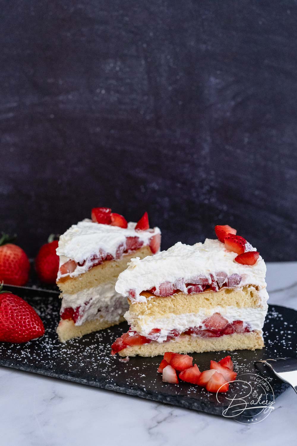 Zwei Tortenstücke der Erdbeer-Sahne-Torte, dekoriert mit Erdbeerstücken auf dunklem Schiefer