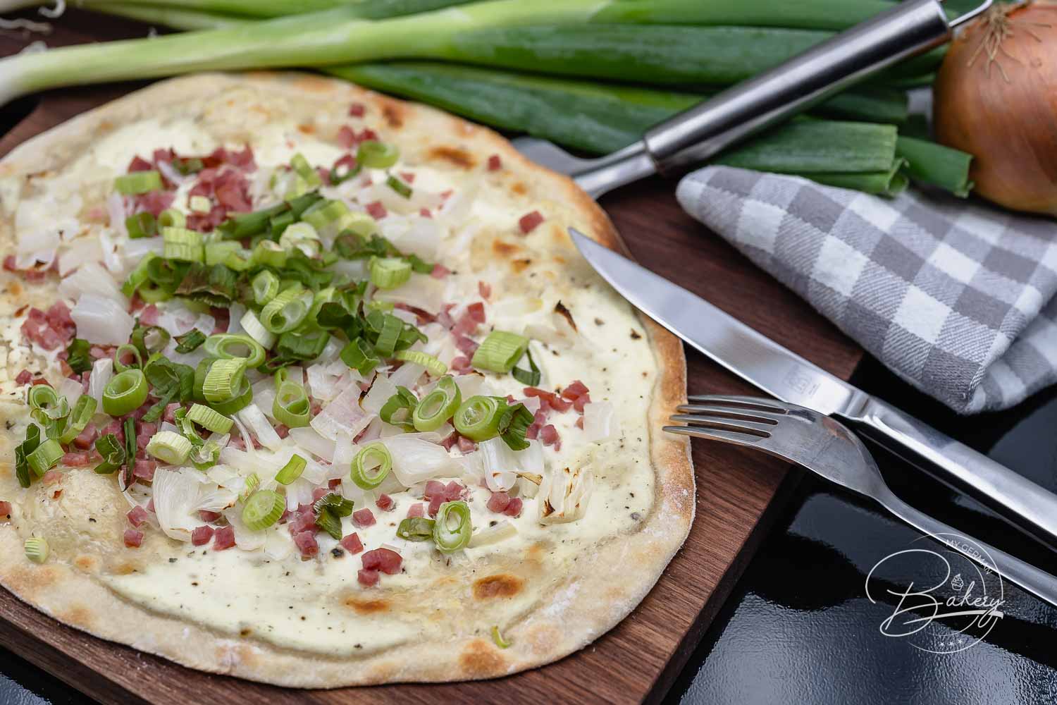 Nahaufnahme vom Flammkuchen mit Speckwürfeln, Zwiebeln und Frühlingszwiebeln, daneben Besteck und kariertes Tuch
