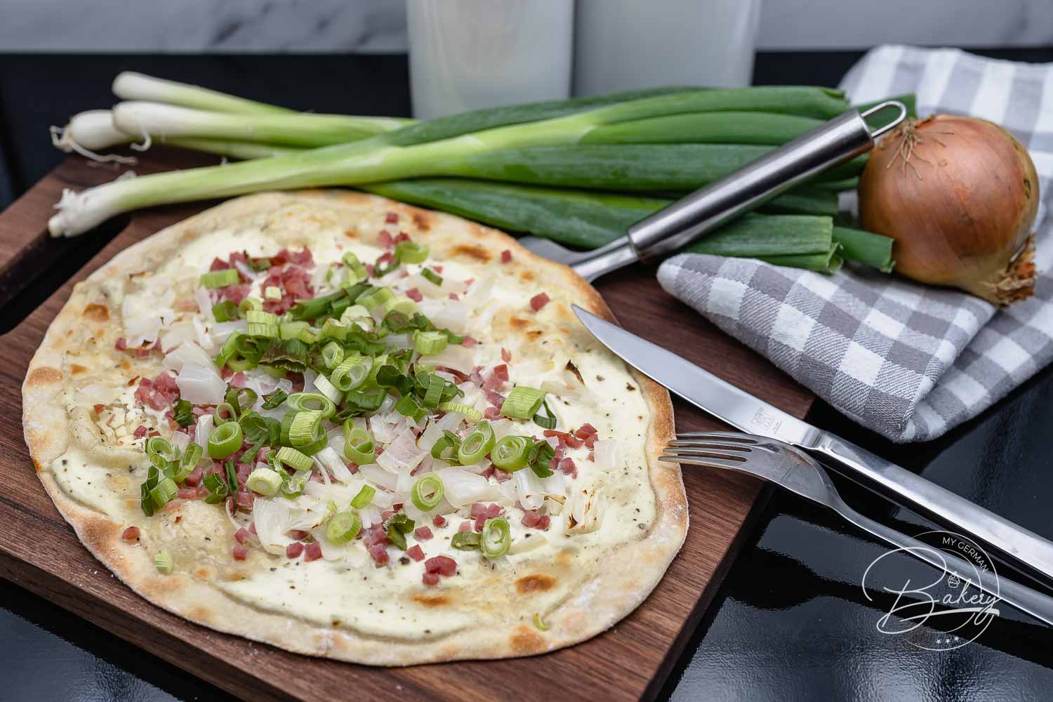 Ganzer Elsässer Flammkuchen mit Speck, Zwiebeln und Frühlingszwiebeln auf Holzbrett, dahinter Lauchzwiebeln und Messer