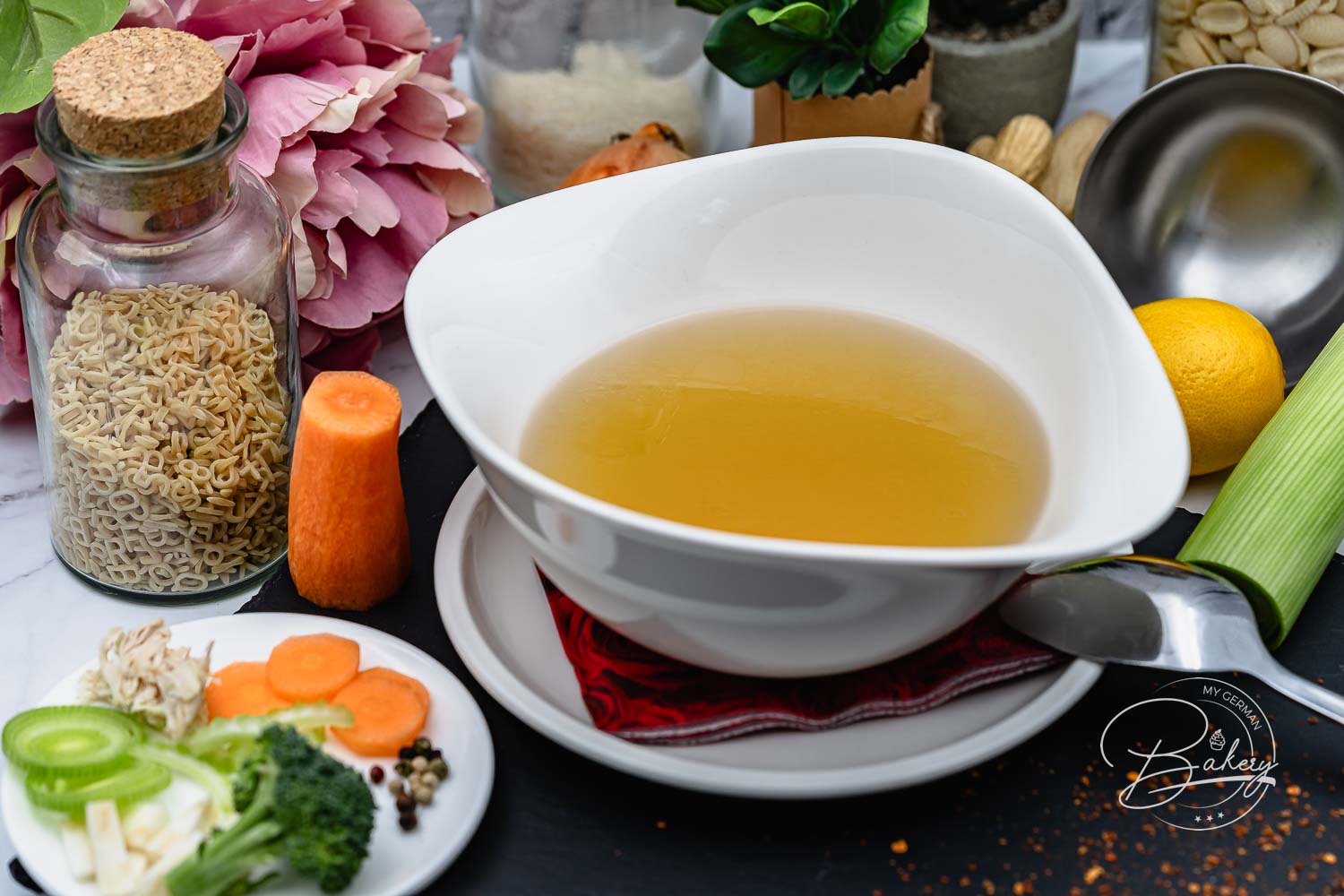 Weiße Schale mit klarer Hühnersuppe-Brühe, umgeben von Nudelglas, Karotte, Gemüseteller und Blumen