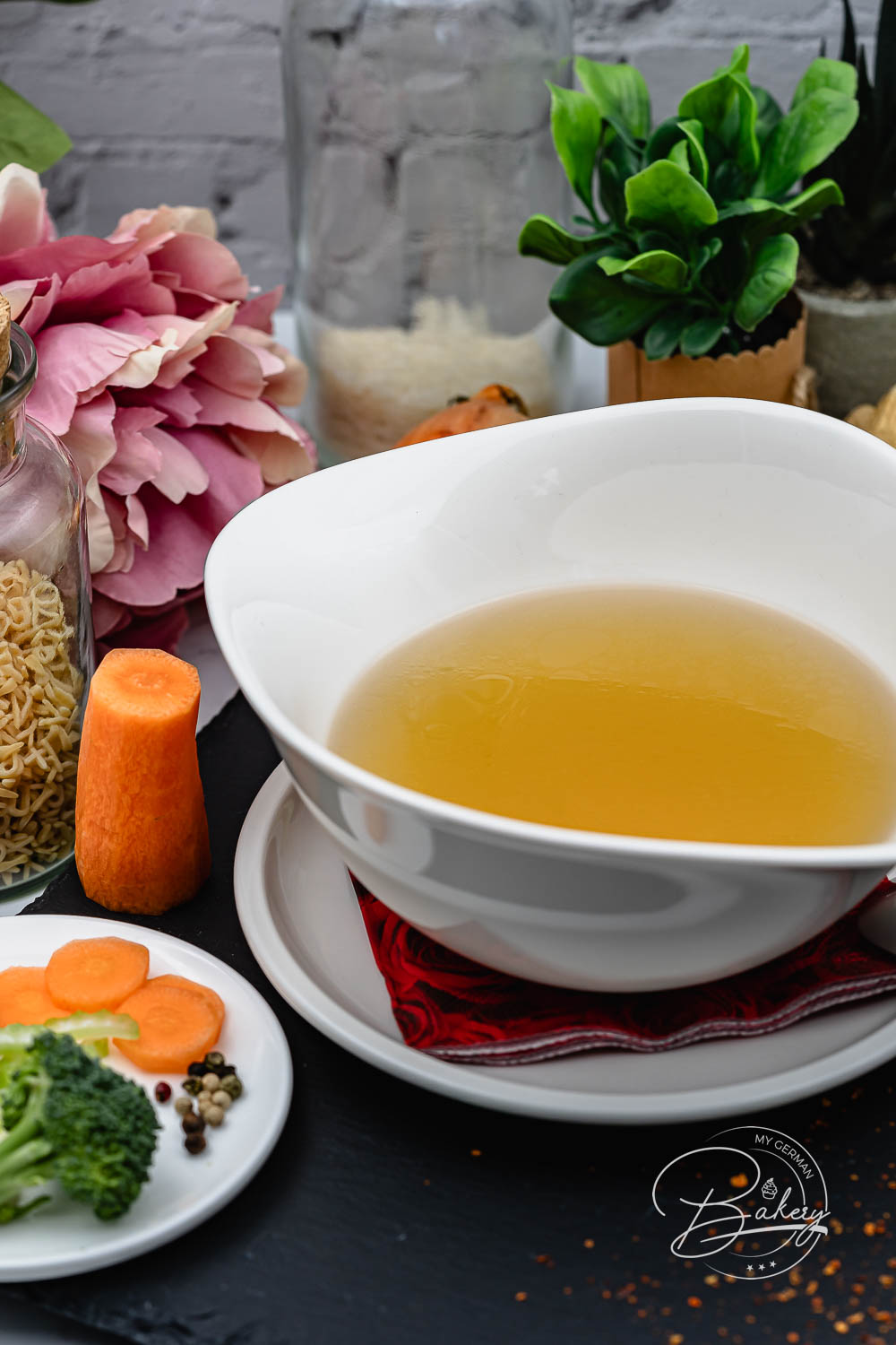 Klare goldene Hühnersuppe-Brühe in weißer Schale, dekoriert mit Karotte, Gemüseteller, Nudeln und rosa Blumen