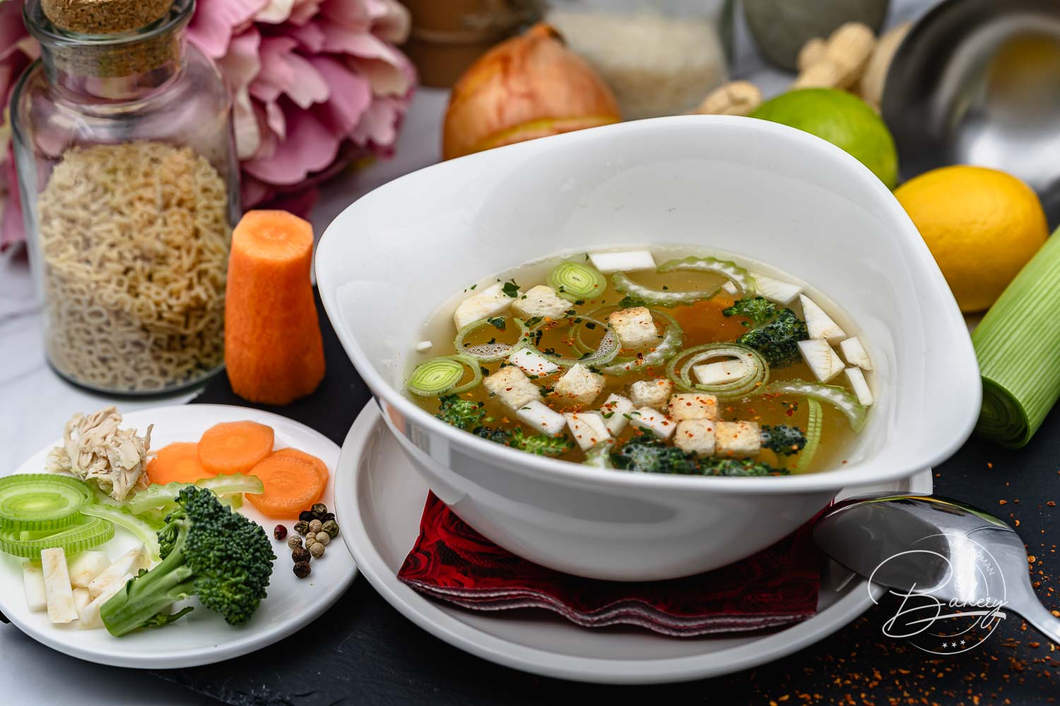 Fertige klassische Hühnersuppe mit Hähnchen, Brokkoli und Lauch in weißer Schale, dazu Gemüseteller