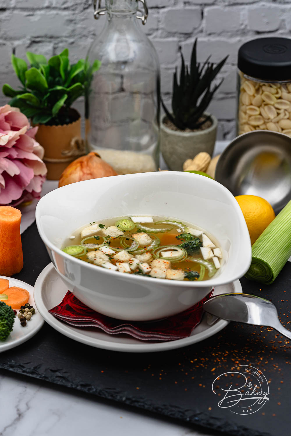 Weiße Schale klassische Hühnersuppe mit Hähnchen, Lauch und Brokkoli, daneben Glasflasche und Nudelglas