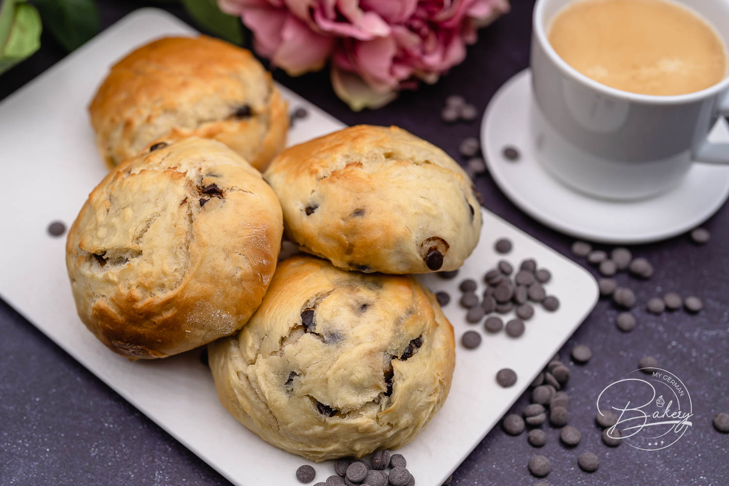 Vier goldbraune Schokobrötchen auf weißer Platte mit Schokodrops, Kaffeetasse und rosa Blüten