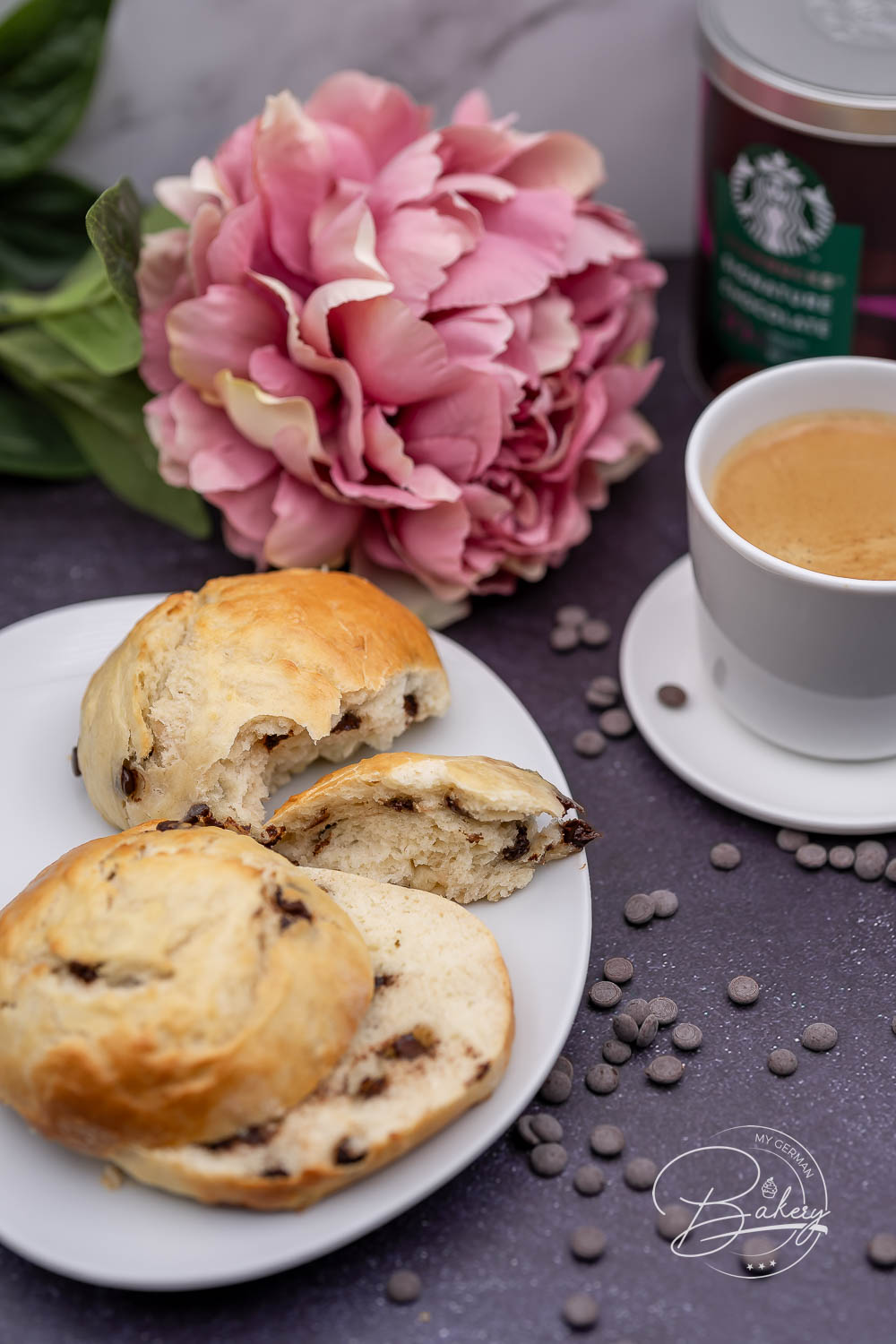 Aufgeschnittenes Schokobrötchen mit Schokostückchen auf weißem Teller neben Espresso und rosa Blüte