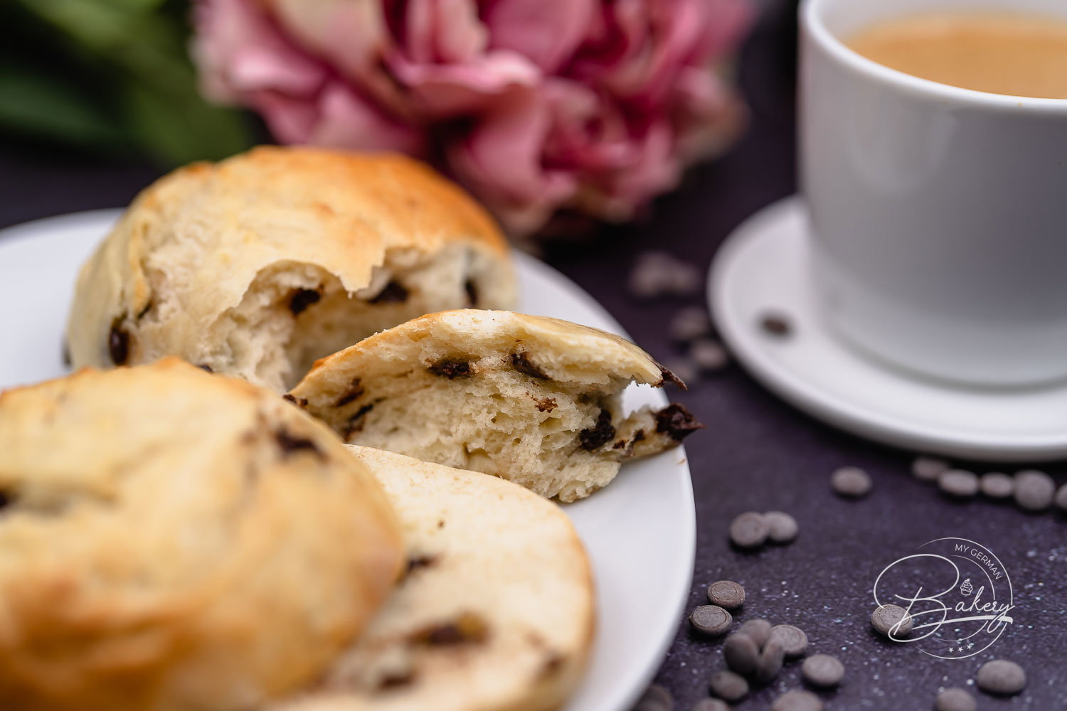 Aufgerissenes Schokobrötchen mit weicher Krume und Schokostückchen auf Teller neben Kaffee und Blüten