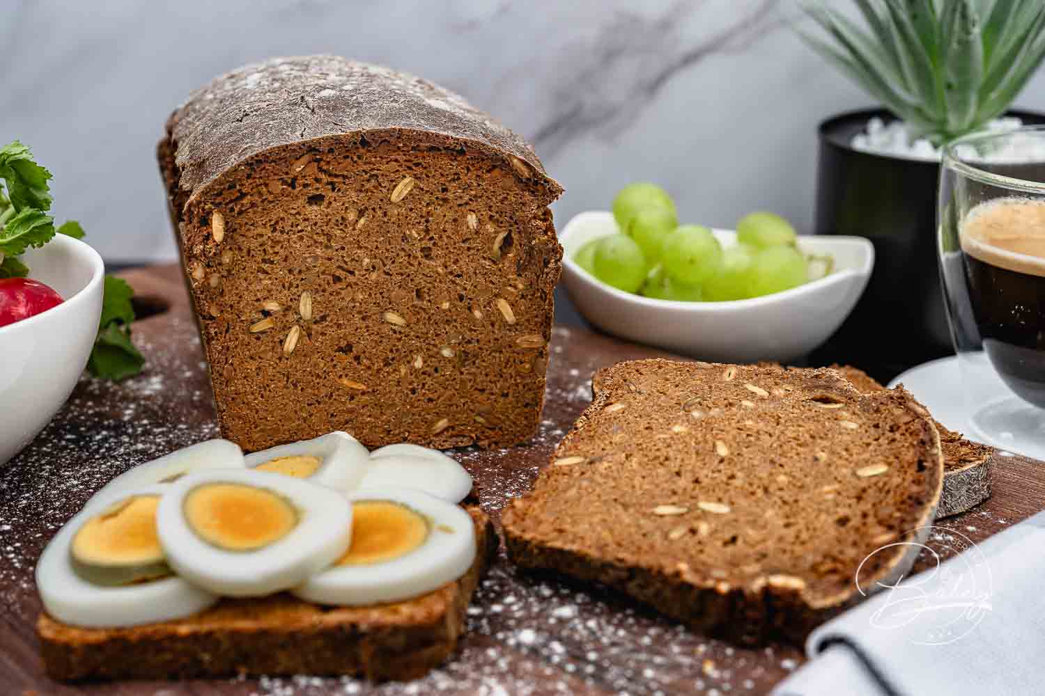 Dunkler Vollkornbrot-Laib auf Holzbrett, davor eine Scheibe mit Ei, Trauben und Espresso – Schwarzbrot Rezept