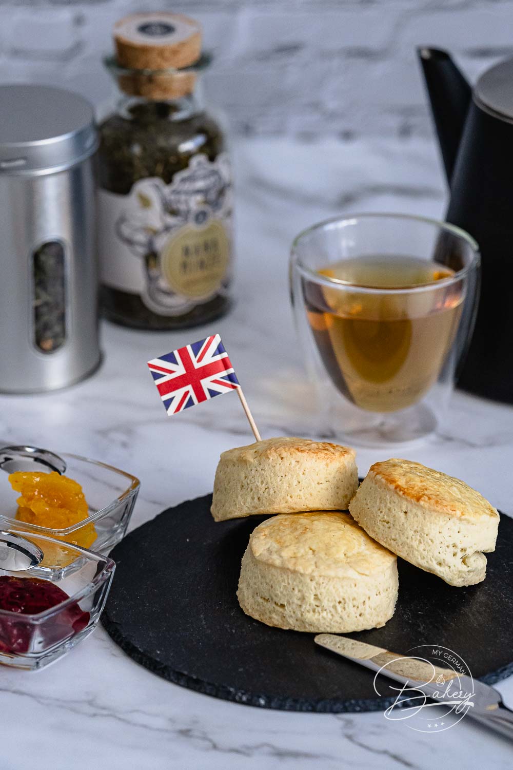 Drei goldbraune Scones auf Schieferplatte, dahinter ein Glas Tee und Teedose