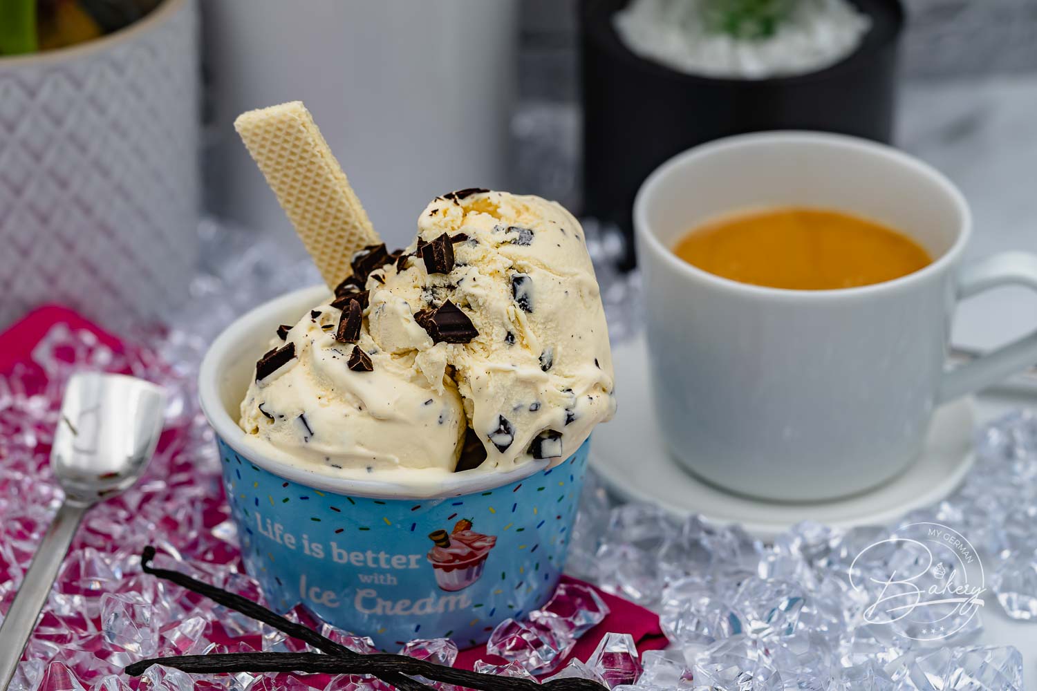 Blauer Eisbecher mit cremigem Stracciatella-Eis, Schokoflocken und Waffelröllchen auf Crushed Ice, Vanilleschoten daneben