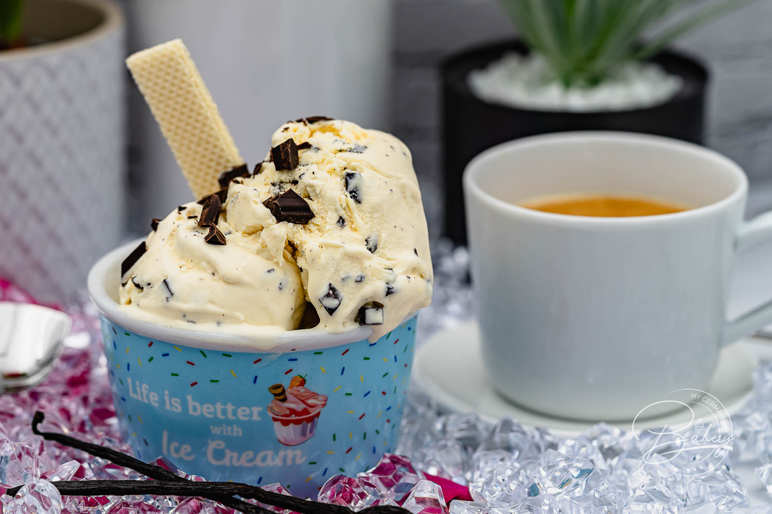 Becher Stracciatella-Eis mit Waffel und Schokoflocken, Espressotasse und Pflanze dahinter, Vanilleschoten auf Crushed Ice
