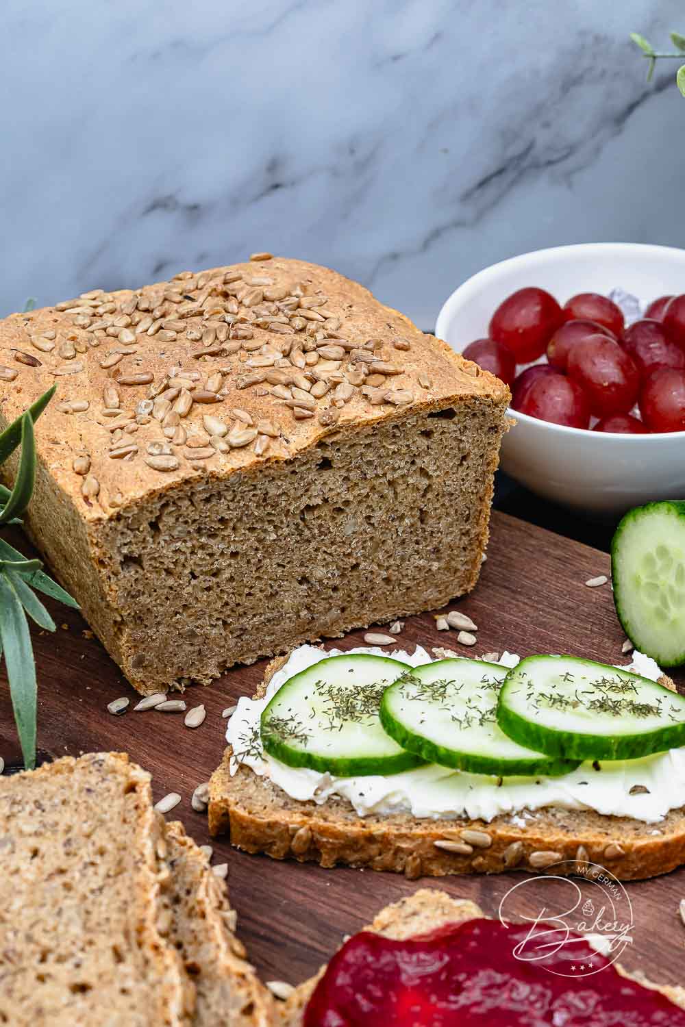 Angeschnittenes Dinkel-Vollkornbrot mit Sonnenblumenkernen neben Weintrauben und einer Scheibe mit Gurke