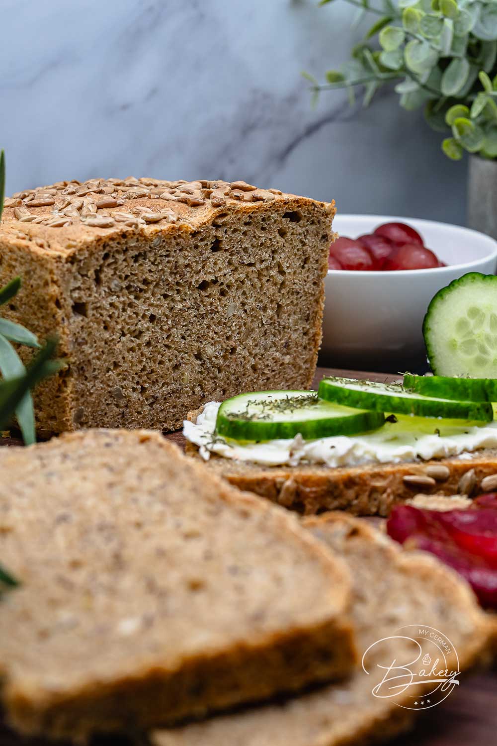 Angeschnittenes Dinkel-Vollkornbrot zeigt die saftige Krume, dazu Scheiben mit Frischkäse und Gurke