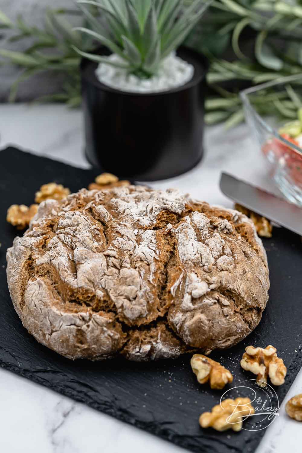 Runder rustikaler Vollkornbrot-Laib mit bemehlter Kruste und Walnüssen auf dunkler Schieferplatte