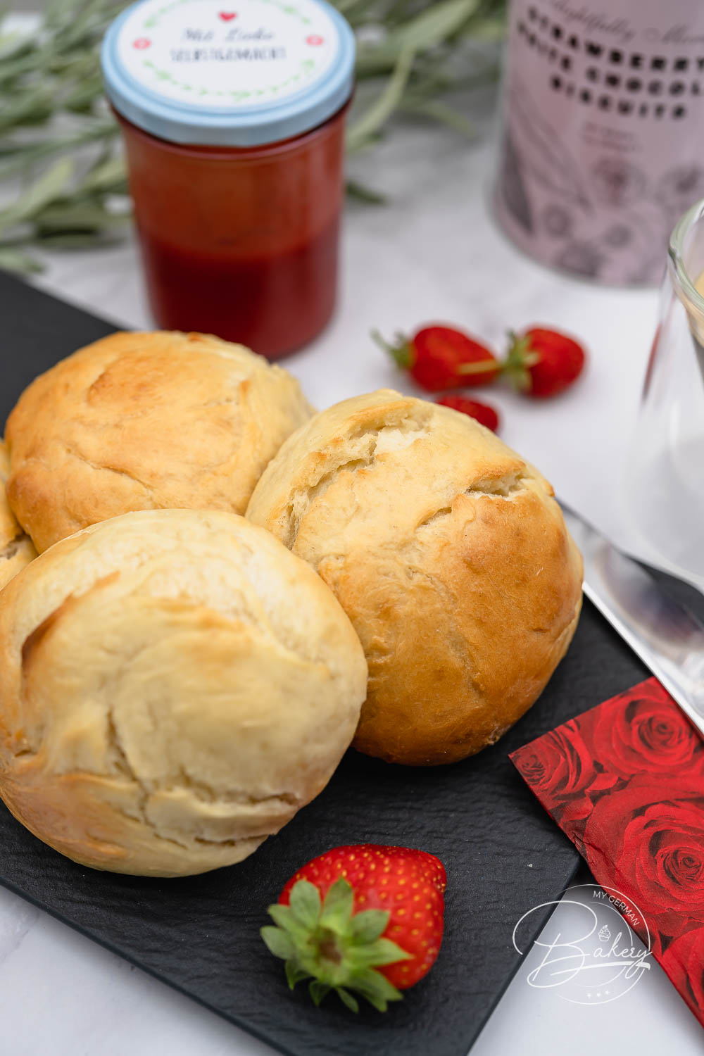 Mehrere Milchbrötchen auf Schiefer mit selbstgemachter Marmelade, frischen Erdbeeren und Rosenserviette