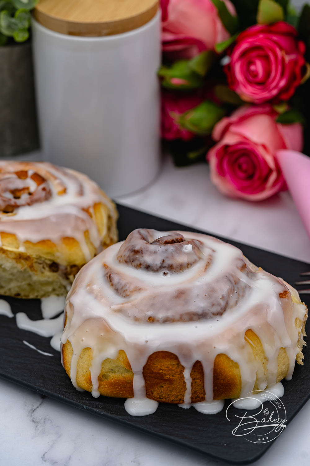 Zwei glasierte Zimtschnecken auf Schiefer, im Hintergrund Rosenstrauß und Vorratsdose