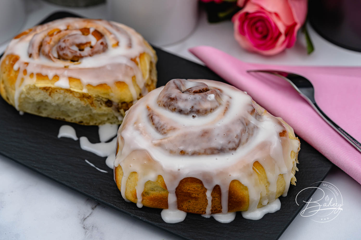 Zwei große Zimtschnecken mit Zuckerguss auf schwarzer Schieferplatte, dahinter Rosen
