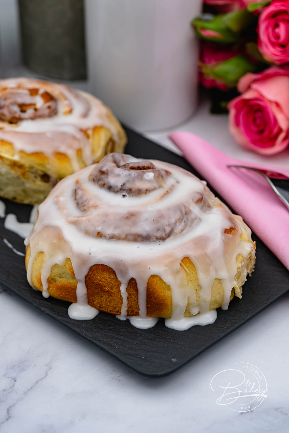 Cinnamon Roll mit fließendem Zuckerguss im Hochformat, rosa Serviette daneben
